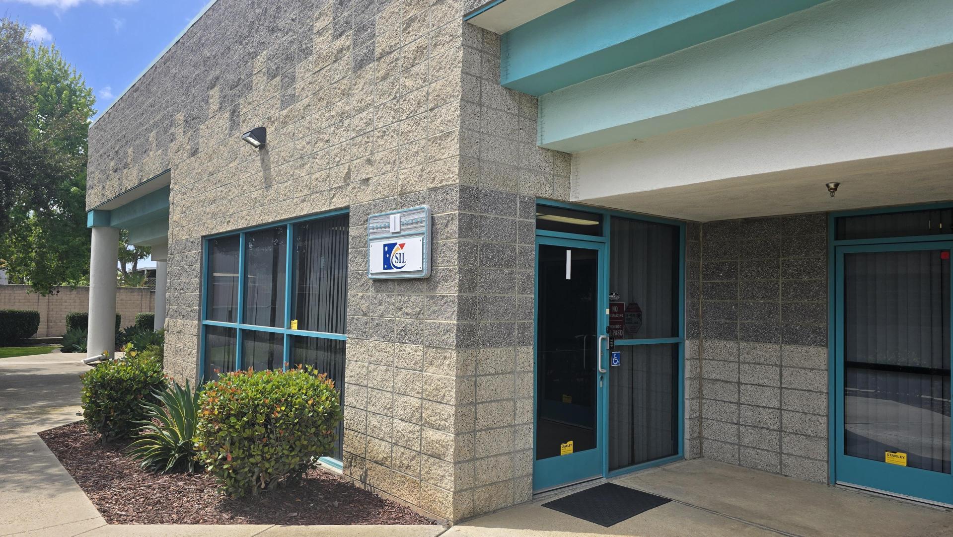 Building exterior with large windows and a sign reading "SIL" above the entrance. Landscaping includes bushes and plants